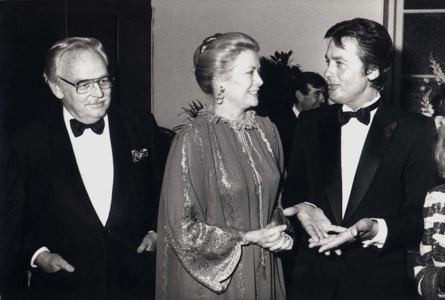 Ez a fotó szintén a korai '80-as években, a Barnum című musical európai premierjén készült, a párizsi Cirque d’Hiver színházban. Grace Kelly egy díszes, hímzett estélyiben, mosolyogva beszélget a legendás francia színésszel, Alain Delonnal, mellette pedig férje, III. Rainier herceg áll. A kép tökéletesen visszaadja a korszak hangulatát – Grace Kelly pedig természetes méltósággal illeszkedett ebbe a világba.