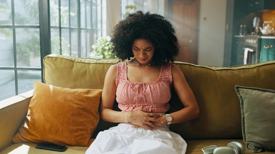 Woman, pain and sofa with stomach ache for indigestion, gastrointestinal disease or reflux at home. Sick, female person or belly cramp with nausea, illness or menstruation for period cycle at house