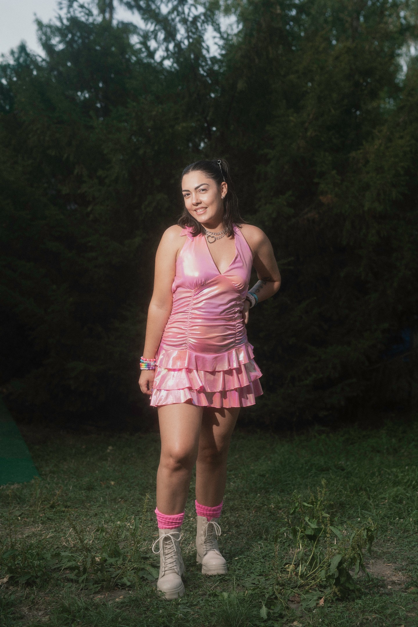 A fényes, metálos rózsaszín anyag egyszerre játékos és figyelemfelkeltő. A ráncolt felsőrész és a fodros miniszoknya együtt teremti meg a csajos, bulikész hangulatot, míg a bakancs és a zokni vagány kontrasztot ad, megtörve a nőies vonalat.