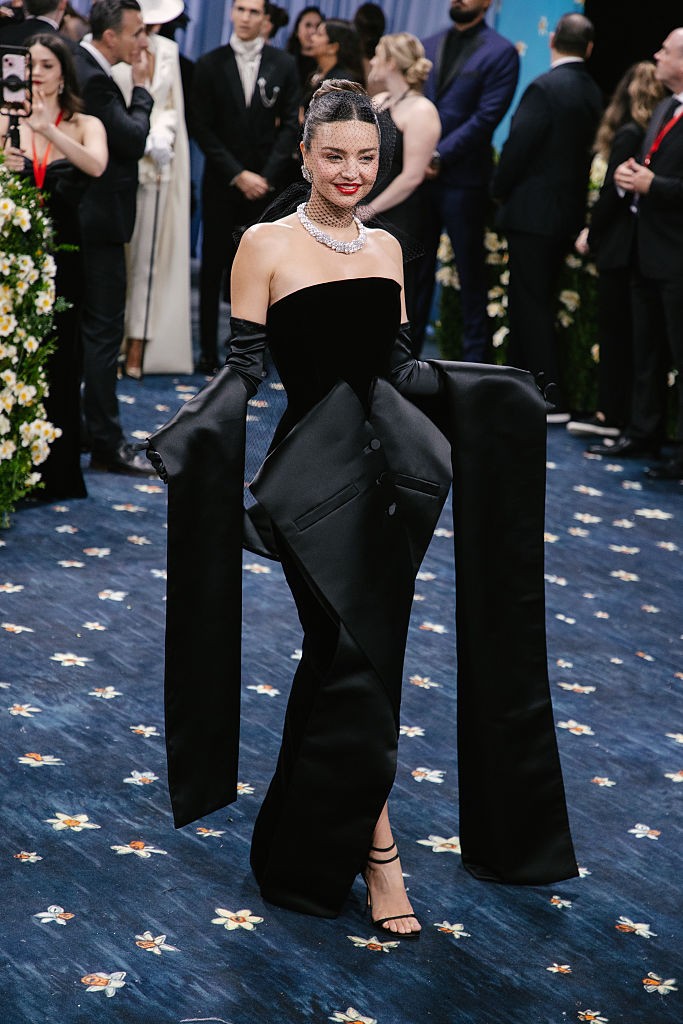 Miranda Kerr – Dior: Miranda Kerr megjelenése a Met-gálán a klasszikus elegancia és a kreatív szabászat tökéletes fúziója volt. A fekete bársony estélyi ruha sziluettje egyszerre idézte meg az old-school glamour és a tailoring világát: a ruha elején zakóként keresztbe hajtott rész és az abból kialakuló, drámai, földig érő ujjak a férfiöltöny ikonikus elemeit értelmezték újra nőies, szoborszerű formában. A gyöngyhálós nyaklánc, a kifinomult konty és a vörös rúzs igazi filmcsillag-hangulatot teremtett. Ez a megjelenés minden részletében a „Tailored For You” szellemiségét tükrözte – finoman, de határozottan.