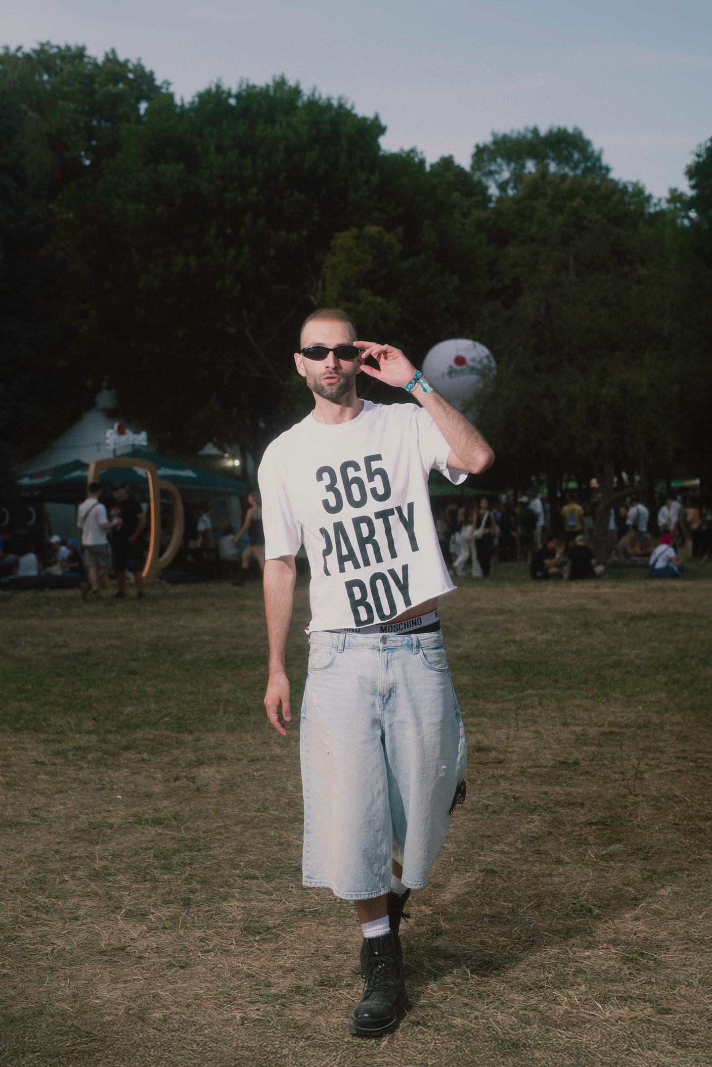 Ez az összeállítás erősen a 2000-es évek elejének Y2K–inspirált, laza utcai stílusát idézi. A bő szárú farmer bermuda és a feliratos póló a korabeli hiphop- és gördeszkás divat tipikus elemei, míg a bakancs egy kissé grunge hatású. A kilógó Moschino alsónadrág gumipántja a 2000-es évek „logómánia” trendjének felel meg. 