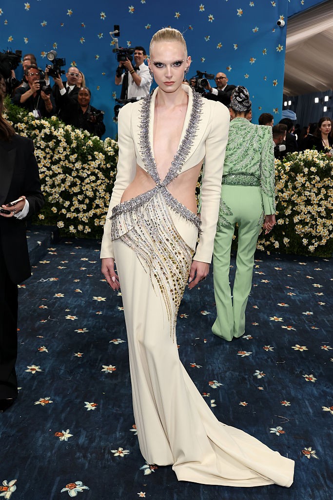 Alex Consani megjelenése egyszerre volt megdöbbentő és lenyűgöző. Az elefántcsontszínű ruha látványos kivágásai szinte semmit nem hagytak a képzeletre, a mellkast pedig egy mélyen szabott, ékköves szegély emelte ki. A csillogó részletek és a szigorú, hátrafogott haj tökéletes egyensúlyt teremtettek a drámai hatás és a high fashion között. 