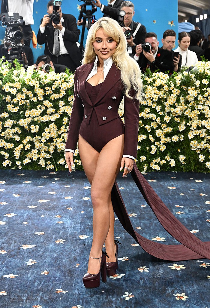 Sabrina Carpenter nadrág nélkül érkezett, és ezzel gyakorlatilag meg is nyerte a Met-gála legmerészebb belépője címet. A bordó, frakkot idéző body egyszerre játszik rá a férfias szabásvonalakra és a nőies idomokra, miközben a fenékvillantás határait feszegeti. Az öltönyfelső gombolása, a fehér gallér és a nyaklánc csak még inkább kiemeli a játékosságot: mintha egy szexi stílusparódiát látnánk egy nagyon is tudatos csomagolásban. A látványos platformcipő és a hajzuhatag pedig már csak hab a tortán!