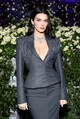 Kendall Jenner ezúttal egy látszólag visszafogottabb szettben érkezett, de ez csak a felszín. A szürke kosztüm precízen szabott vállai és karcsúsított derékvonala megidézi a klasszikus férfisziluettet, miközben a mély dekoltázs rögtön elárulja, hogy itt bizony szó sincs szigorú irodai viseletről. Pontosan ez a konzervatív forma és a meztelenség finom utalása közötti vibrálás teszi ennyire izgalmassá ezt a megjelenést. 