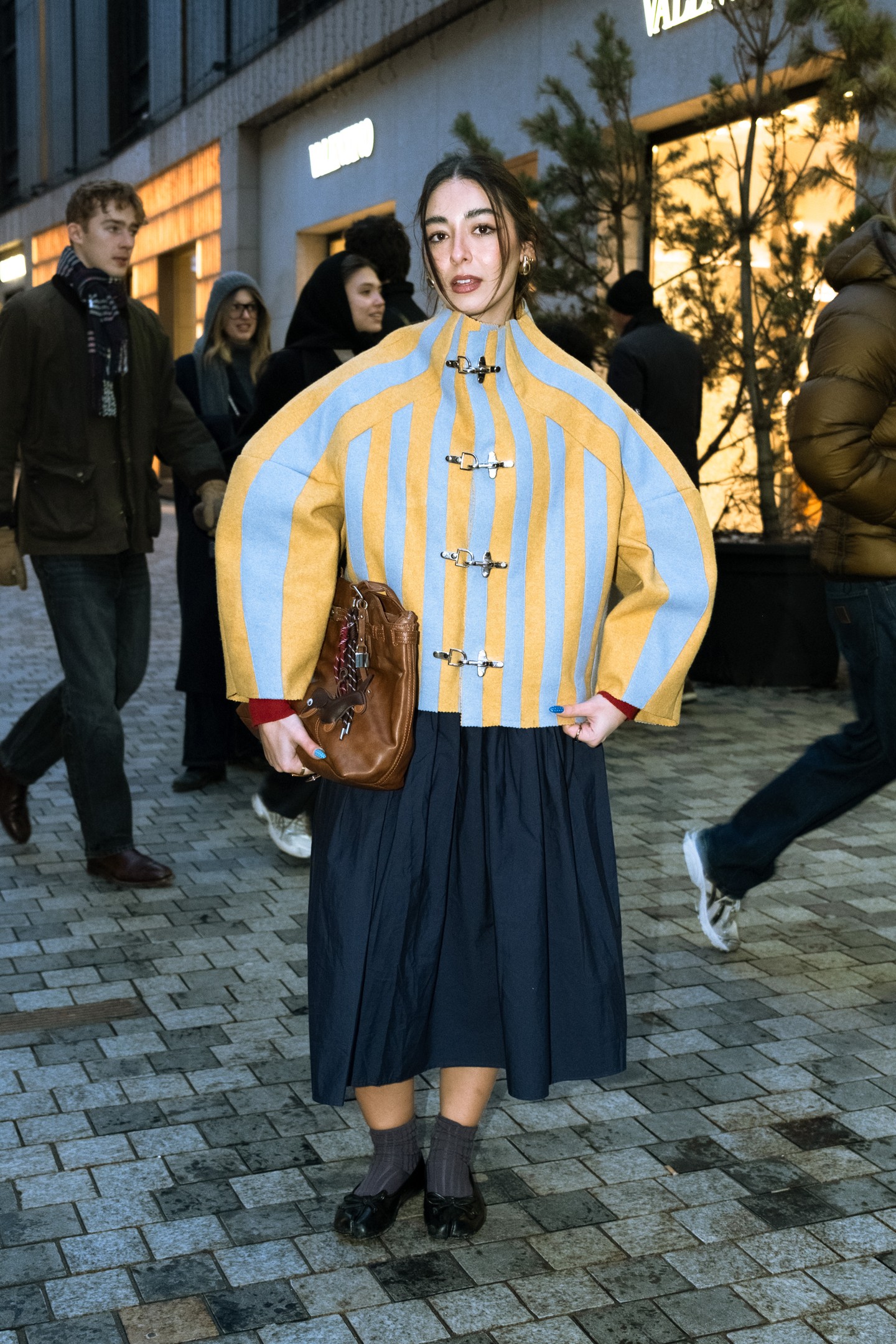 A szoborszerű, csatos kabát adja a szett fókuszát, miközben a lágy szoknya és a finom kiegészítők ellensúlyozzák az erős formákat.