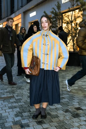 A szoborszerű, csatos kabát adja a szett fókuszát, miközben a lágy szoknya és a finom kiegészítők ellensúlyozzák az erős formákat.