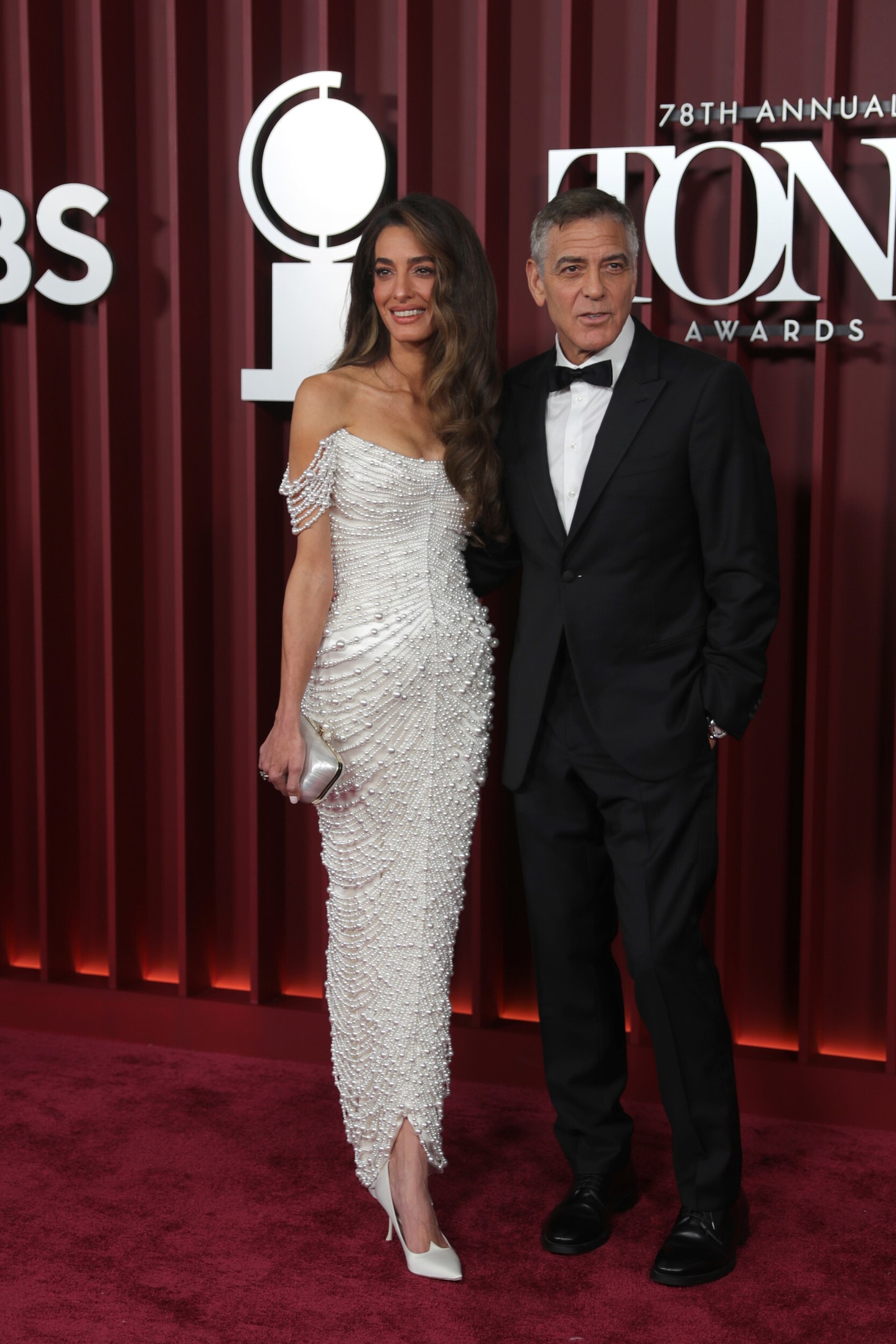 Amal Clooney az idei Tony-gálára egy testhez simuló, hófehér Tamara Ralph kreációt viselt fűzőszerű, vállvillantós felsőrésszel, amit különböző méretű gyöngyök borítottak. 