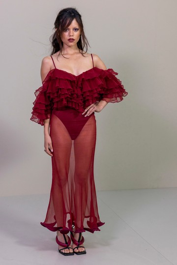 Jenna Ortega a 2025-ös párizsi divathéten egy áttetsző, fodrokkal díszített, bordó Givenchy pucérruhában tündökölt. 