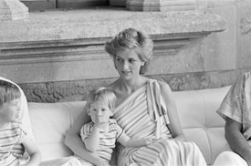 Diana hercegné egy elegáns, váll nélküli ruhában pihen a mallorcai nyaraláson, miközben ölében tartja a hüvelykujját szopizó, még totyogós Harry herceget. 