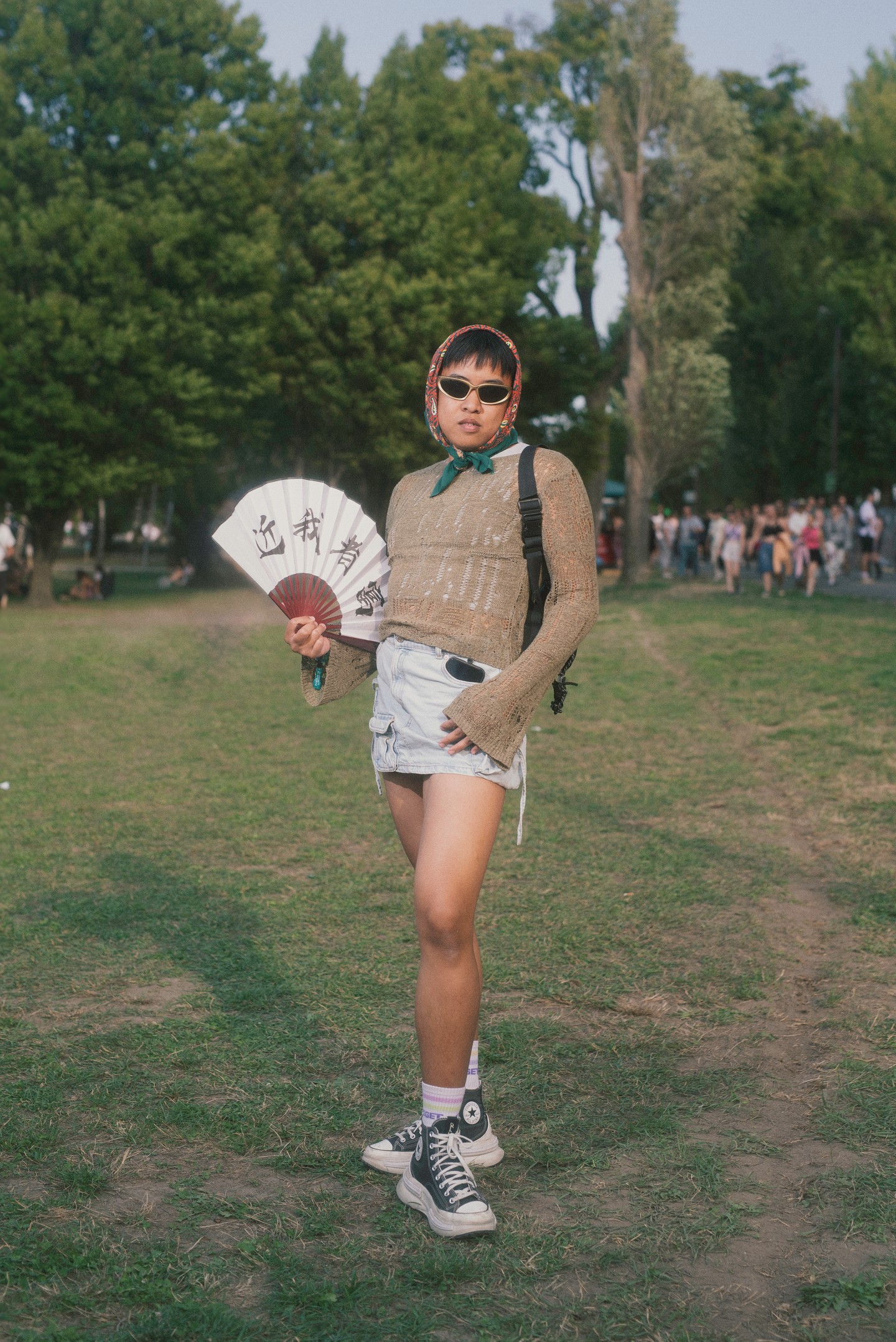 Ez az outfit remek példája annak, milyen felszabadult és kreatív lehet az öltözködés a Sziget Fesztiválon. Viselője laza, mégis tudatosan összeválogatott darabokat kombinál: a szakadt, áttetsző pulóver és a rövid, húzott farmerszoknya kontrasztja játékosan hat, miközben a fejkendő, a retró napszemüveg és a kézben tartott japán legyező különleges, bohém hangulatot kölcsönöz a megjelenésnek.