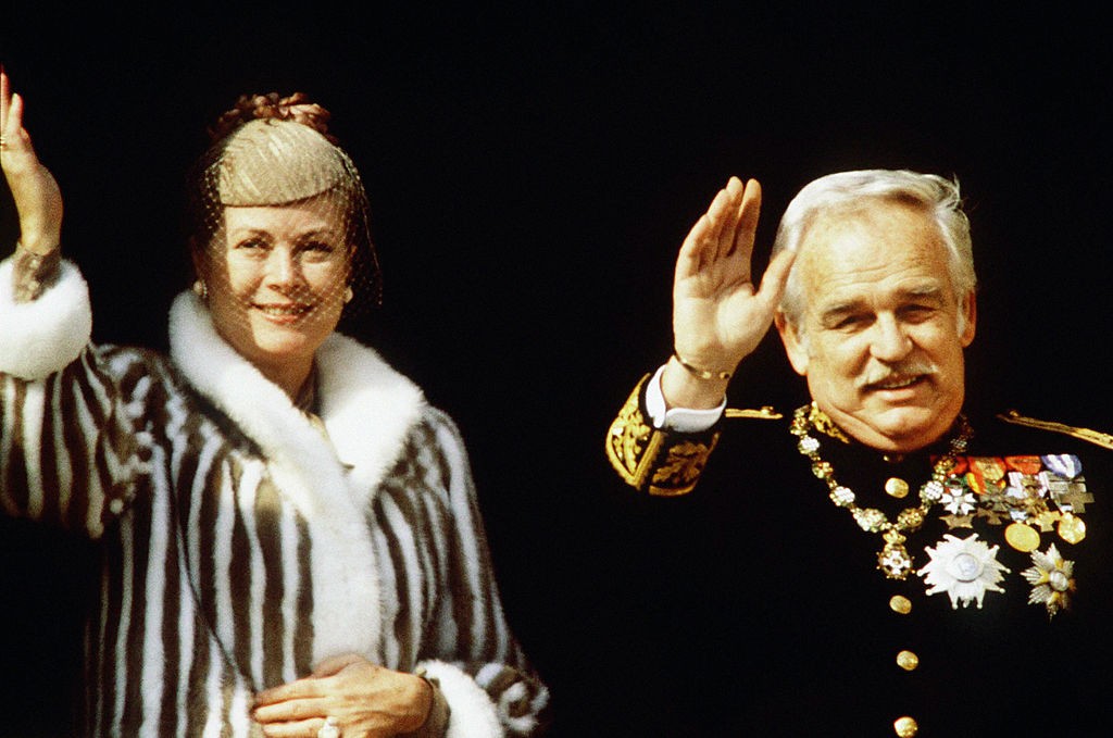 Grace Kelly és III. Rainier herceg, amint a nemzeti ünnep alkalmából integetnek a monacói palota erkélyéről. A hercegné különleges szőrmekabátban áll férje mellett – mosolyog, miközben megőrzi azt a méltóságteljes viselkedést, ami mindig is jellemezte. A kép egyszerre hivatalos és emberi, tökéletesen összefoglalja közös éveik elegáns lezárását.