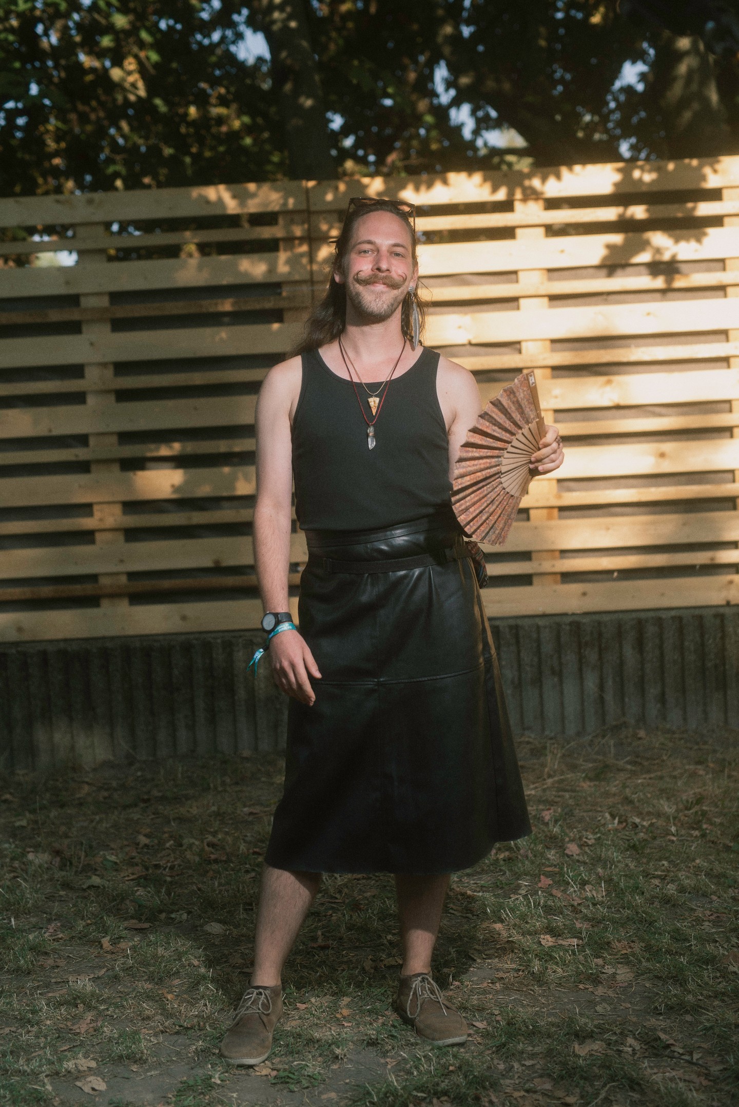 Egyedi stílusjegyek és fesztiválhangulat találkozik ebben az összeállításban. A fekete ujjatlan felső és a térdig érő bőrszoknya minimalista, mégis karakteres párost alkot, amelyet a kiegészítők boho és alternatív irányba terelnek. A kézben tartott legyező és a barna mokaszinok játékosan oldják a szett rockos, fekete alapját.